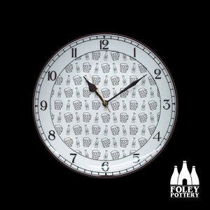 May include: A white ceramic clock with black numbers and hands. The clock face has a pattern of black beer mugs. The clock is framed with a black rim and has a black and white logo that says "FOLEY POTTERY" at the bottom.