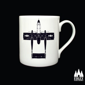 May include: A white ceramic mug with a black silhouette of an A-10 Thunderbolt II aircraft printed on the front. The aircraft is facing downwards with its wings spread out.