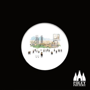 May include: A white ceramic coaster with a hand-drawn illustration of a mining town. The illustration features a mine shaft, buildings, and a group of people in yellow vests. The text "FLONARD" is written on a sign in the foreground.