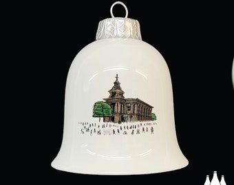 May include: White ceramic Christmas ornament shaped like a bell with a hand-drawn illustration of a building with a clock tower and people walking in front of it. The building is labeled "BUCKLEY TOWN HALL".