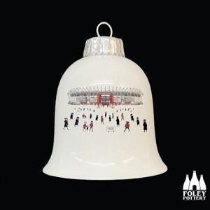May include: White ceramic Christmas ornament shaped like a bell with a black and white illustration of a stadium and people. The stadium is red and white and has the words "Parkhead Stadium" printed on it.