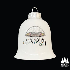 May include: White ceramic bell-shaped Christmas ornament featuring a detailed illustration of Wembley Stadium with people walking towards it. The ornament has a silver cap and a loop for hanging. The words "Wembley Stadium" are visible on a sign.