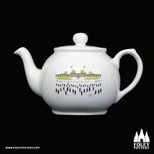 May include: White teapot with a black and white illustration of a building with flags and people in front of it. The teapot has a round body and a handle on the right side.