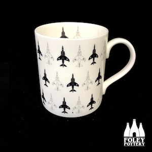 May include: A white ceramic mug with a black and white pattern of jet fighter planes. The mug has a black handle and the words "FOLEY POTTERY" are printed on the bottom.