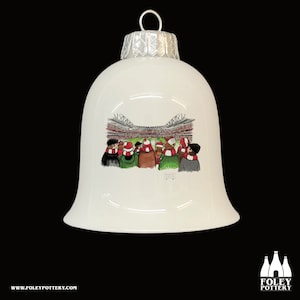May include: White ceramic Christmas ornament shaped like a bell with a red and white stadium scene and a group of people wearing scarves and hats. The ornament has a silver top.