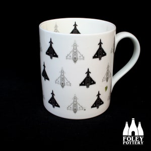 May include: White ceramic mug with a black and white pattern of fighter jets. The mug has a black handle and a white interior.