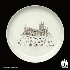 Puede incluir: Un plato de cerámica blanco con una ilustración en blanco y negro de la Catedral de Gloucester y una multitud de personas frente a ella. El texto "GLOUCESTER CATHEDRAL" está impreso en el plato.