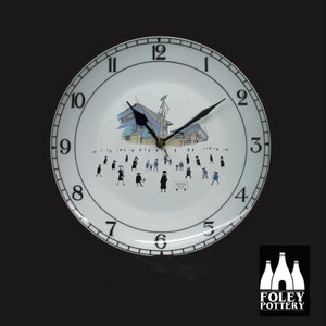 May include: White ceramic clock with black numbers and hands. The clock face features a black and white illustration of a stadium with people walking around it. The text "Portman Road" is visible on the clock face. The clock is made by Foley Pottery.