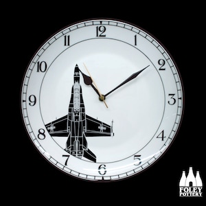 May include: A white ceramic clock with black Roman numeral hour markers and black hands. A black silhouette of a fighter jet is in the center of the clock face.