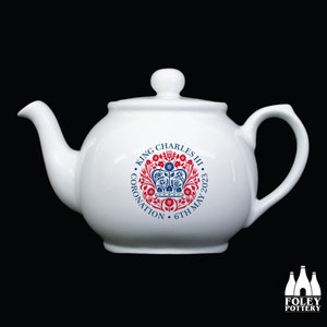 May include: White ceramic teapot with a lid and handle. The teapot features a circular emblem with the text "KING CHARLES III CORONATION 6TH MAY 2023" in red and blue. A commemorative item for the coronation.