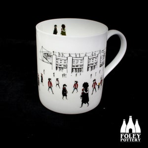 Puede incluir: Taza de cerámica blanca con una ilustración en blanco y negro de un estadio y personas caminando hacia él. La ilustración incluye algunas personas con camisetas rojas y blancas. La taza tiene un asa negra.
