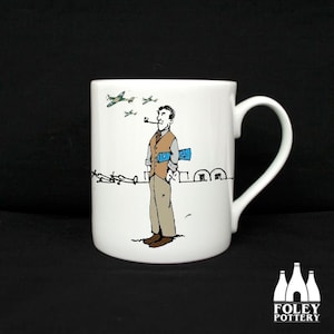 Op de afbeelding: Witte keramische mok met een cartoonillustratie van een man in een bruin vest en blauwe sjaal, die een pijp vasthoudt en voor een prikkeldraadhek staat. Er vliegen kleine groene vliegtuigen in de lucht boven hem. De mok heeft een zwart handvat.