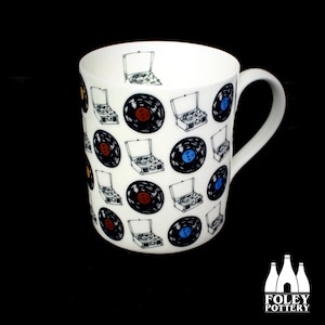 May include: White ceramic mug with a black and white pattern of vintage record players and vinyl records. The records have red, blue, and yellow accents.