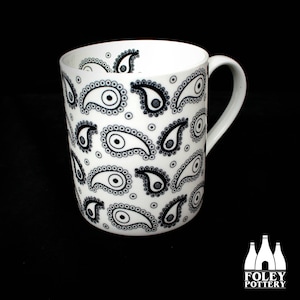 May include: White ceramic mug with a black and white paisley pattern. The mug has a handle and the words "FOLEY POTTERY" are printed on the bottom.
