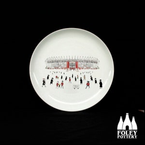 May include: White ceramic plate with a black and white illustration of a stadium with people walking towards it. The stadium is red and white. The text "ELLAND ROAD STADIUM" is printed on the plate.