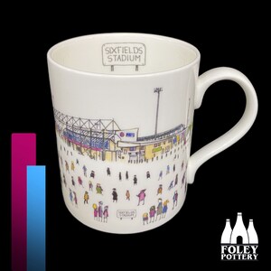 Peut inclure: Une tasse en céramique blanche avec une illustration en noir et blanc d'un stade, avec les mots "Sixfields Stadium" sur une pancarte en haut. L'illustration comprend de nombreuses petites personnes de différentes couleurs. La tasse a une poignée blanche.