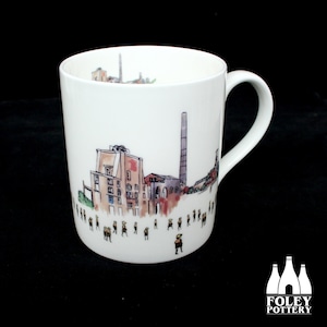 May include: White ceramic mug with a hand-drawn illustration of a factory and people in front of it. The mug has a black handle and the words "FOLEY POTTERY" are printed on the bottom.