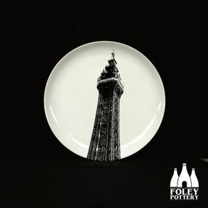 May include: A white ceramic plate featuring a black and white silhouette of a tall tower. The tower has a pointed top and numerous windows. The plate is set against a black backdrop.