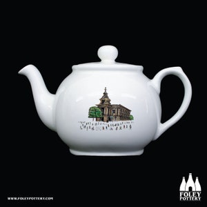 May include: White ceramic teapot with a black and white illustration of a building with a clock tower and people walking in front of it.