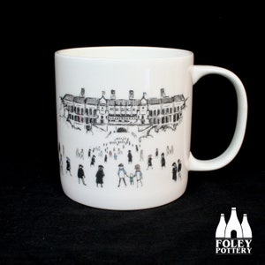 May include: White ceramic mug with a black and white illustration of a building with people walking in front of it. The building has a large archway and several windows. The people are all wearing hats and coats. The mug has a black handle.