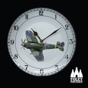 Op de afbeelding: Een witte keramische klok met een zwarte wijzerplaat en zwarte wijzers. De klok heeft een groen en bruin vliegtuig met rode, witte en blauwe markeringen. Het vliegtuig is een Supermarine Spitfire met het registratienummer "UO4U". De klok is gemaakt door Foley Pottery.