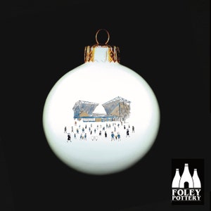 Peut inclure: Boule de Noël blanche avec une illustration dessinée à la main d'un stade avec des personnes devant. La boule est décorée d'une garniture dorée. Le texte "FOLEY POTTERY" est imprimé au bas de la boule.
