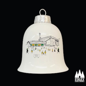 May include: White ceramic Christmas ornament shaped like a bell with a black and white illustration of a stadium with people in front of it. The text "Carrow Road" is on a sign in front of the stadium.