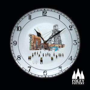 May include: White ceramic clock with a black clock face and black hands. The clock face features a black and white illustration of a coal mine with a group of miners in yellow and black uniforms. The clock face also includes the number 12 and the word "LITTLETON".