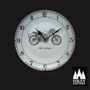 May include: White ceramic clock with black numbers and hands. The clock face features a black and white illustration of a vintage motorcycle with the text "BSA - Goldstar -" underneath. The clock is made by Foley Pottery.