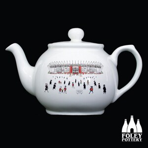 May include: White ceramic teapot with a black and white illustration of a stadium with people in front of it. The stadium is red and white. The text "SUPER SOCCER WEEKEND" is on a sign in front of the stadium.