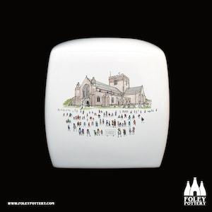 May include: A white ceramic toilet tank lid with a hand-drawn illustration of St. Asaph Cathedral in Wales. The illustration shows the cathedral with a crowd of people in front of it.