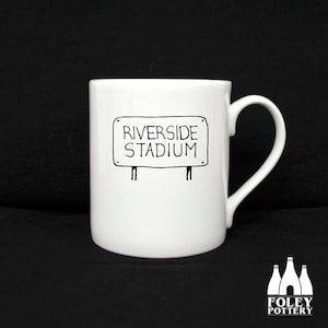 May include: White ceramic mug with a black and white illustration of a sign that reads "Riverside Stadium".