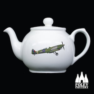 May include: White ceramic teapot with a green and brown airplane design. The airplane has a red, white, and blue roundel on the side. The teapot is made by Foley Pottery.