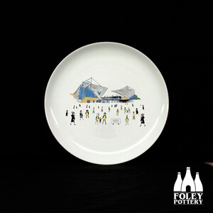 Op de afbeelding: Witte keramische plaat met een stadionillustratie in blauw, geel en grijs. De plaat toont een menigte mensen en een bord met de tekst "KASIM STADIUM". Het Foley Pottery-logo staat rechtsonder.