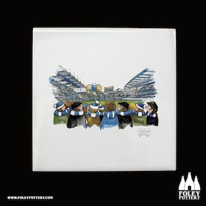 May include: A white ceramic tile with a cartoon illustration of a group of people wearing blue and white scarves, watching a soccer game at Stamford Bridge. The tile has the words "Stamford Bridge" printed on it.