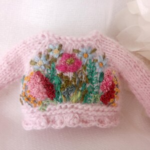 K&ouml;nnte beinhalten: Ein hellrosa Strickpullover mit langen &Auml;rmeln. Der Pullover hat eine bunte Blumenstickerei auf der Vorderseite, darunter rosa, rote, blaue und gr&uuml;ne Blumen. Der untere Saum hat einen gewellten Rand.