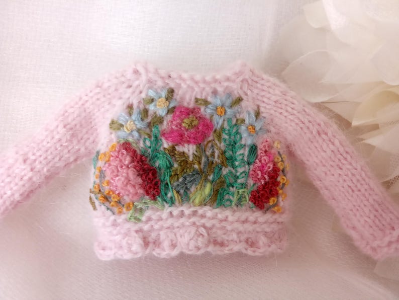 K&ouml;nnte beinhalten: Ein hellrosa Strickpullover mit langen &Auml;rmeln. Der Pullover hat eine bunte Blumenstickerei auf der Vorderseite, darunter rosa, rote, blaue und gr&uuml;ne Blumen. Der untere Saum hat einen gewellten Rand.