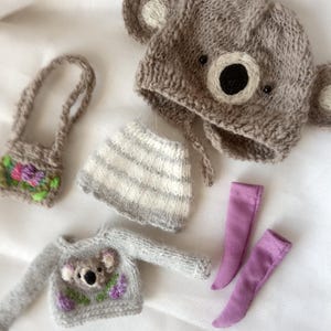 May include: A collection of handmade doll clothes, including a gray koala bear hat, a gray sweater with a koala design, a striped skirt, a small purse, and two lavender-colored socks. The items are made of soft, knitted materials.