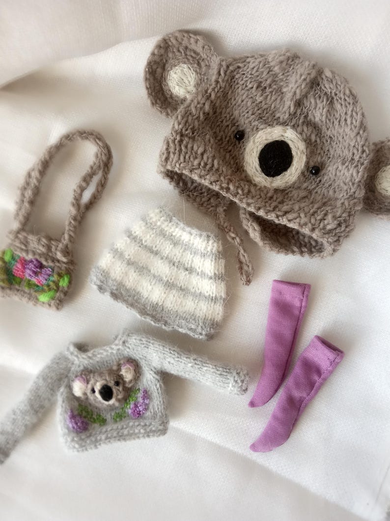 May include: A collection of handmade doll clothes, including a gray koala bear hat, a gray sweater with a koala design, a striped skirt, a small purse, and two lavender-colored socks. The items are made of soft, knitted materials.