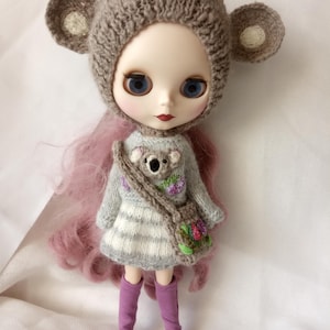 May include: A doll with blue eyes and pink hair wearing a gray knitted dress with a koala design and a matching purse. The doll is wearing a brown bear hat with ears and purple boots. The doll is approximately 12 inches tall.