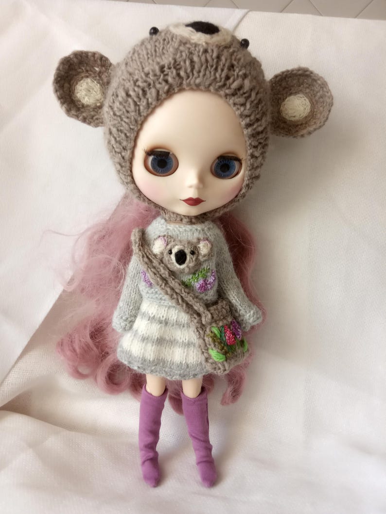 May include: A doll with blue eyes and pink hair wearing a gray knitted dress with a koala design and a matching purse. The doll is wearing a brown bear hat with ears and purple boots. The doll is approximately 12 inches tall.