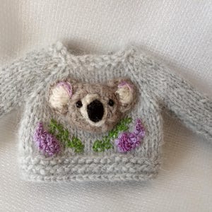 May include: A miniature gray knitted sweater featuring a koala bear design on the front. The koala is brown with white and pink accents, surrounded by purple flowers and green leaves. The sweater has long sleeves.