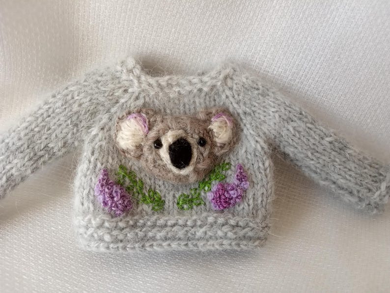 May include: A miniature gray knitted sweater featuring a koala bear design on the front. The koala is brown with white and pink accents, surrounded by purple flowers and green leaves. The sweater has long sleeves.