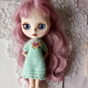 Puede incluir: Una mu&ntilde;eca con el pelo rosa que lleva un vestido de punto verde claro con bordados florales. La mu&ntilde;eca tiene ojos azules y est&aacute; posando con las manos en las caderas.