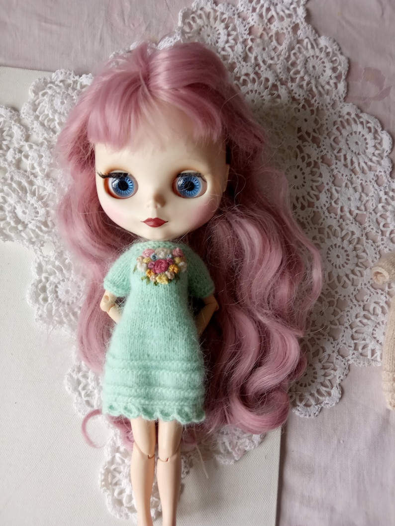 Puede incluir: Una mu&ntilde;eca con el pelo rosa que lleva un vestido de punto verde claro con bordados florales. La mu&ntilde;eca tiene ojos azules y est&aacute; posando con las manos en las caderas.