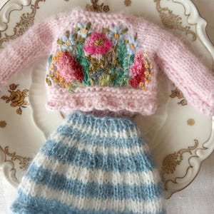 K&ouml;nnte beinhalten: Ein Miniatur-Set aus einem rosa Strickpullover mit Blumenstickerei und einem passenden blau-wei&szlig; gestreiften Rock. Der Pullover hat einen Rundhalsausschnitt und lange &Auml;rmel. Der Rock hat einen gewellten Saum. Das Set wird auf einem dekorativen Teller pr&auml;sentiert.