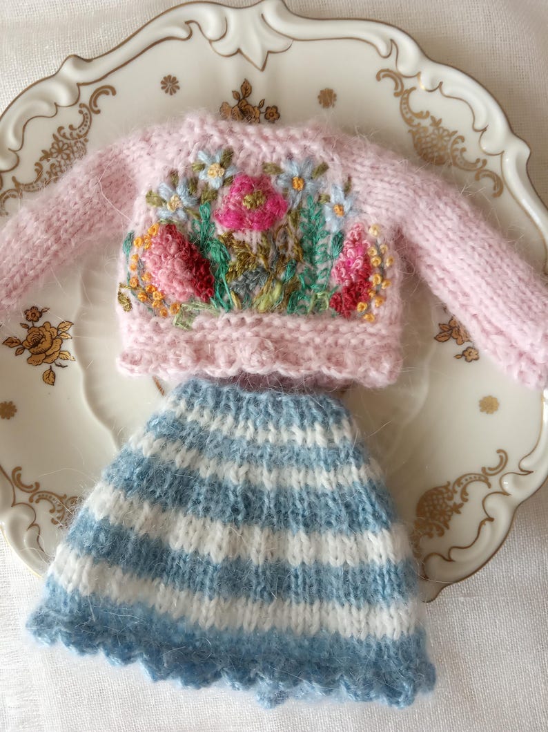 K&ouml;nnte beinhalten: Ein Miniatur-Set aus einem rosa Strickpullover mit Blumenstickerei und einem passenden blau-wei&szlig; gestreiften Rock. Der Pullover hat einen Rundhalsausschnitt und lange &Auml;rmel. Der Rock hat einen gewellten Saum. Das Set wird auf einem dekorativen Teller pr&auml;sentiert.