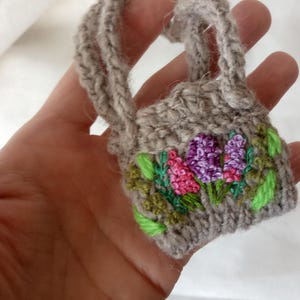 May include: A miniature gray knitted handbag with embroidered flowers in shades of pink and purple, accented by green leaves. The bag has a small handle and is held in a hand.
