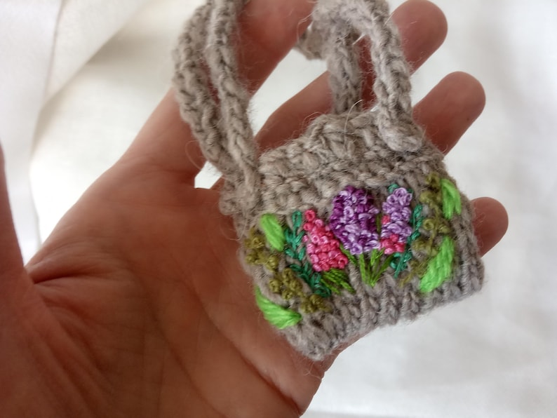 May include: A miniature gray knitted handbag with embroidered flowers in shades of pink and purple, accented by green leaves. The bag has a small handle and is held in a hand.