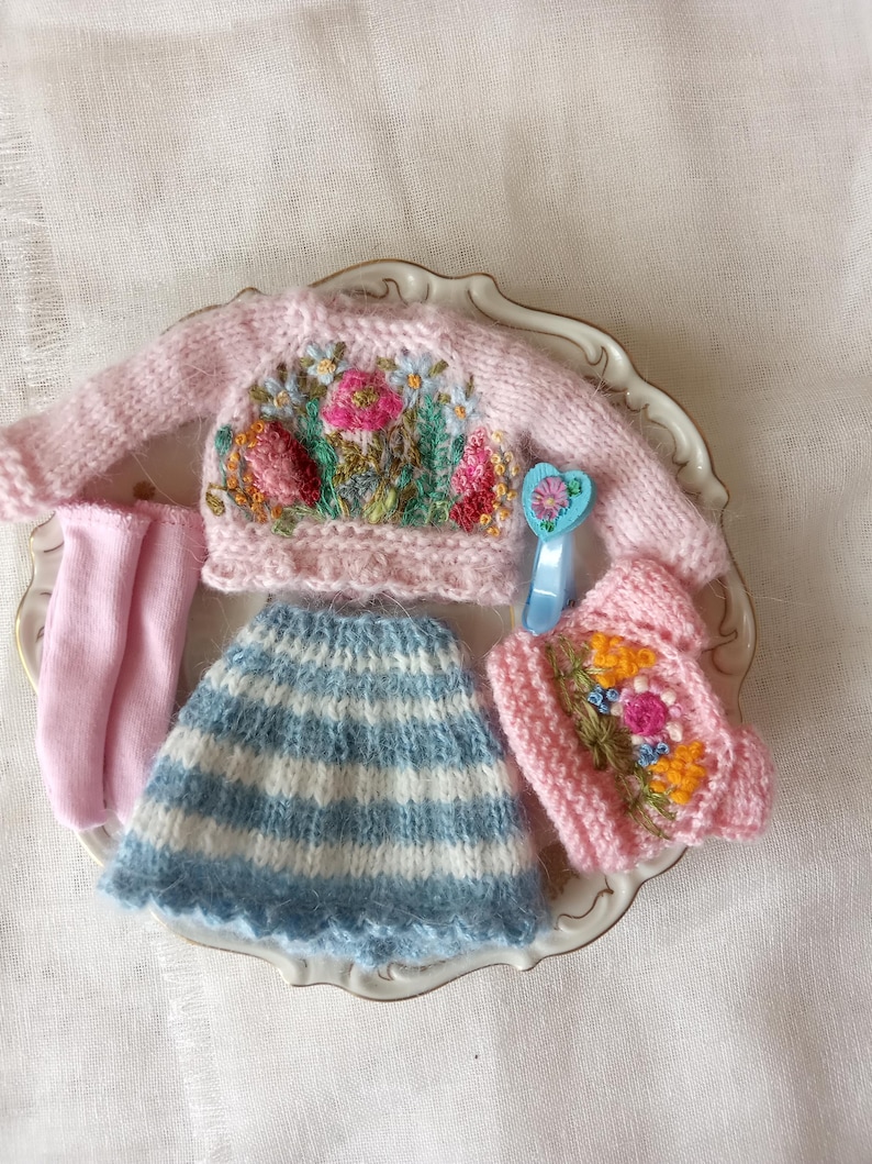 K&ouml;nnte beinhalten: Ein Miniatur-Puppen-Outfit mit einem rosa Pullover mit Blumenstickerei, einem blau-wei&szlig; gestreiften Rock, rosa Socken und einer rosa M&uuml;tze. Das Outfit wird auf einem dekorativen Teller pr&auml;sentiert und zeigt die filigrane Handwerkskunst der handgefertigten Kleidung.